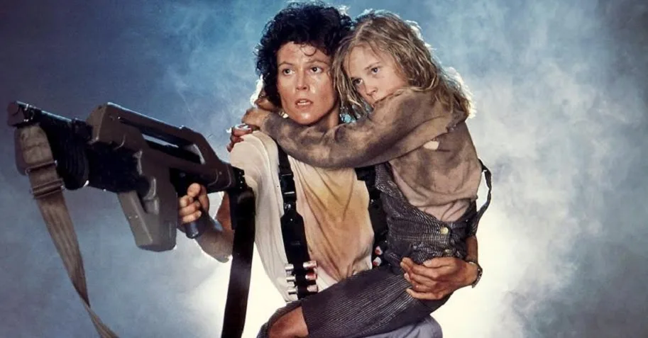 A woman with curly dark hair, holding a large sci-fi weapon, looks alarmed while carrying a young girl in a dirty, muddy…