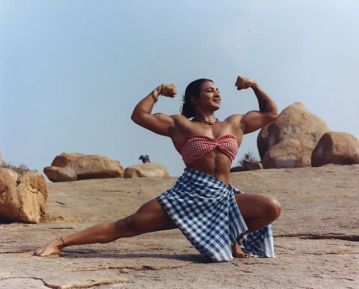 A muscular woman with tattoos poses on rocky terrain, flexing both arms while in a wide-legged squat. She wears a…