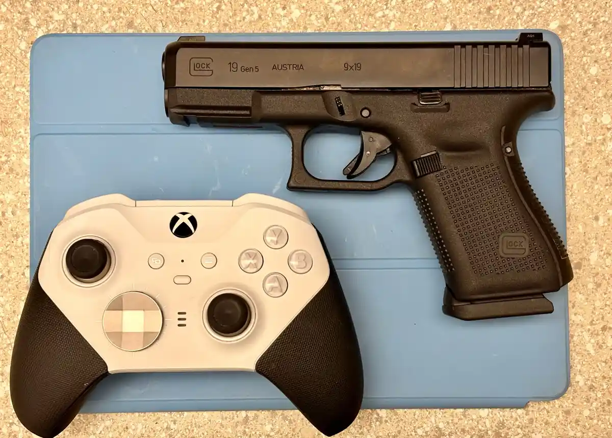 A white and black Xbox controller and a black Glock 19 Gen 5 pistol are placed side by side on a blue plastic surface. The…