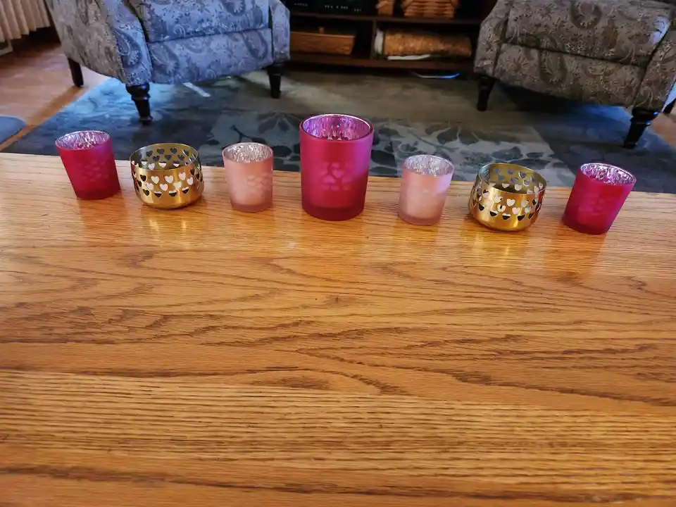 Six decorative candle holders in pink and gold, some with heart cutouts, are arranged in a row on a wooden coffee table in a…