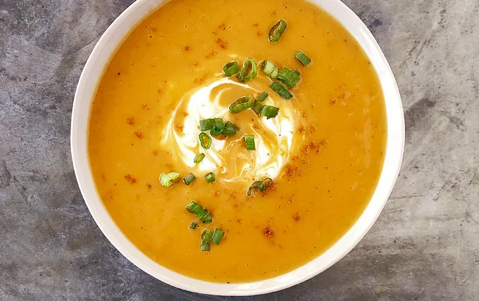 A top-down photo of a white bowl filled with creamy orange soup, garnished with a swirl of white cream and chopped green…