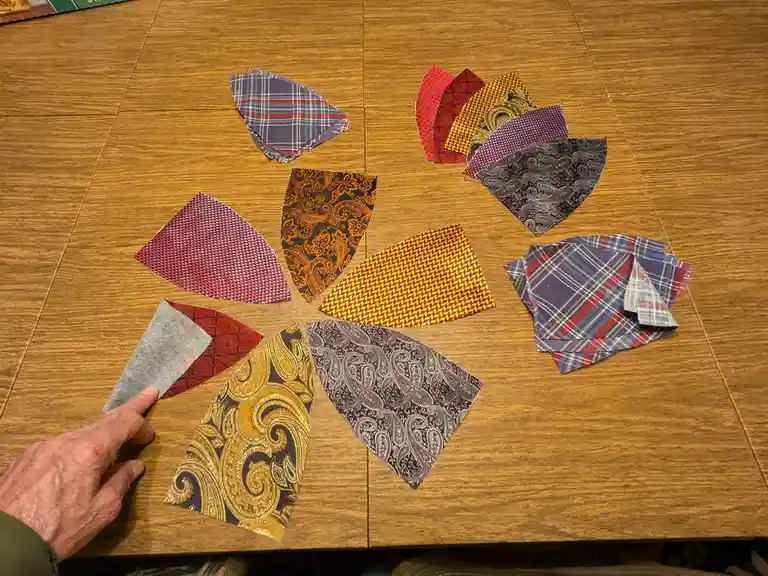 A hand points to a plain gray fabric swatch among a circular arrangement of colorful, patterned tie samples on a wooden…