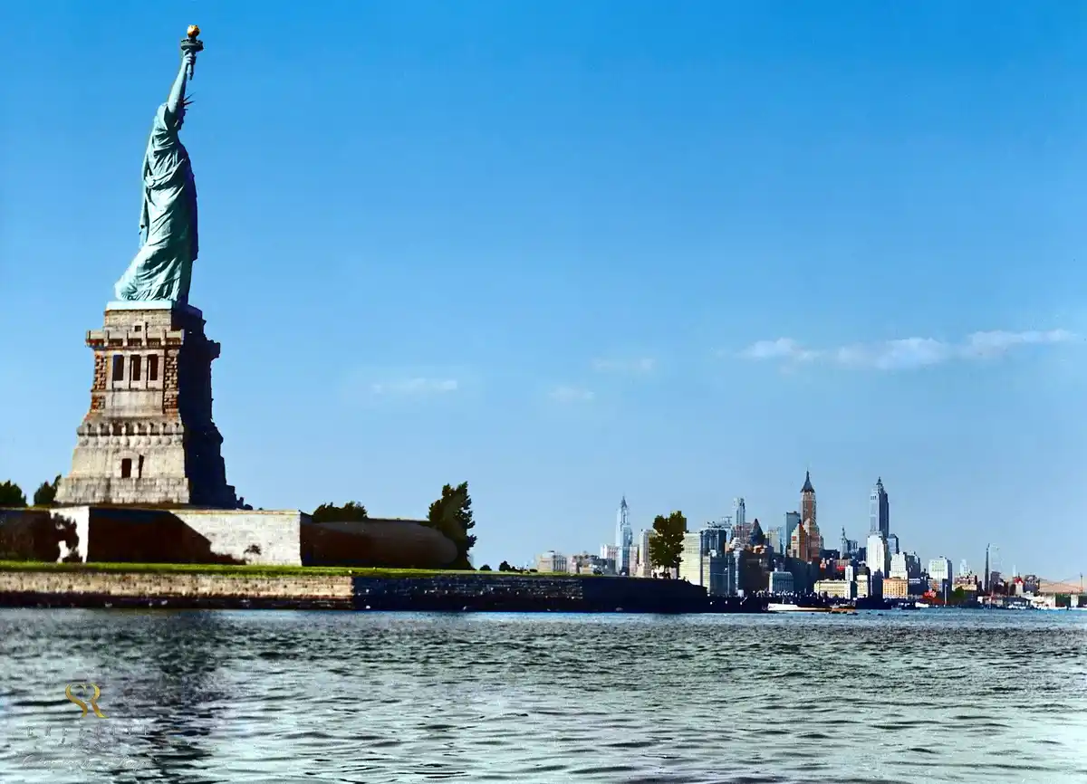 A vintage-style photograph shows the Statue of Liberty standing on its pedestal in New York Harbor, with the Manhattan…