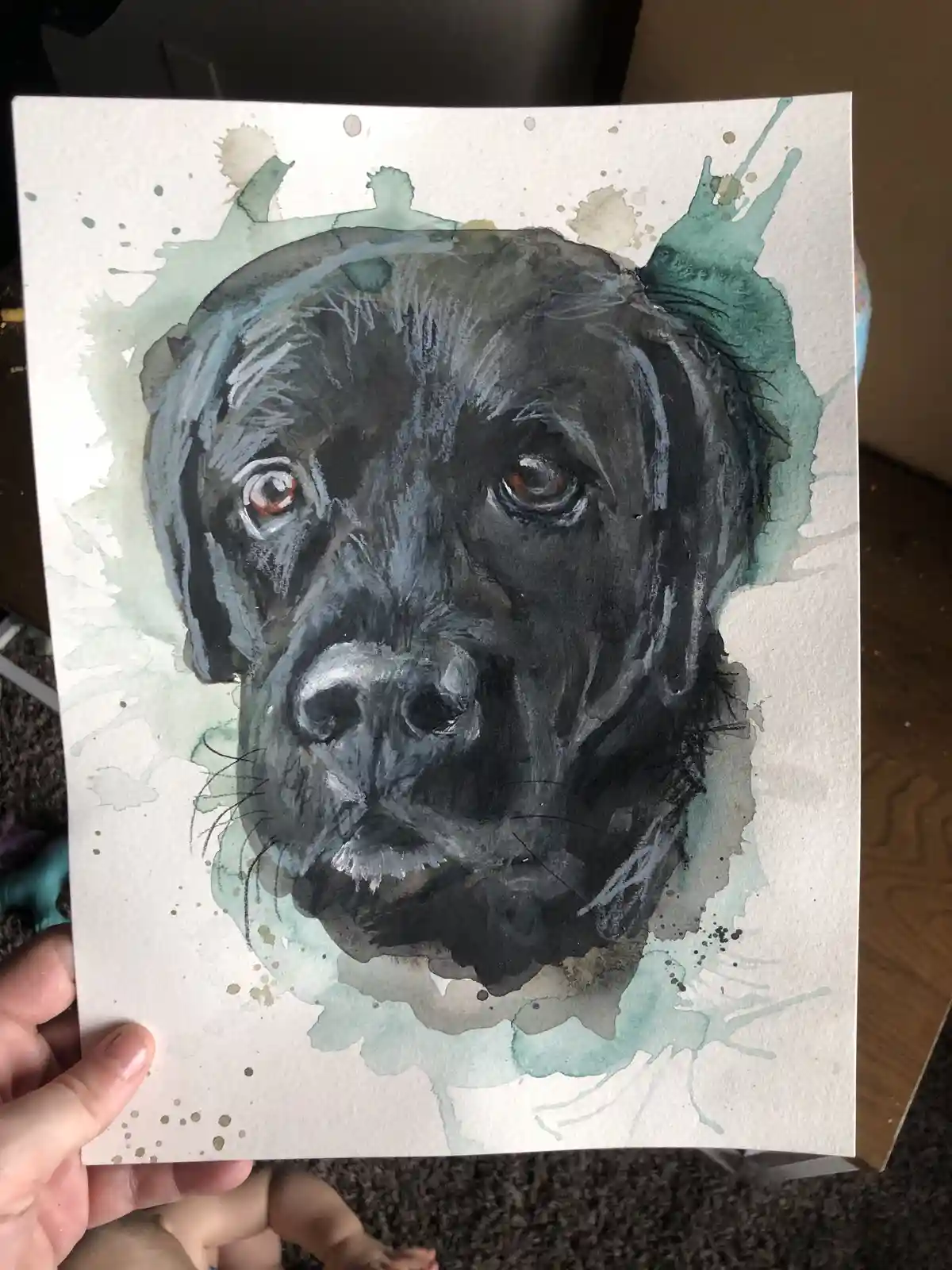 A hand holds up a watercolor painting of a black dog's face with expressive eyes and a serious expression, surrounded by…