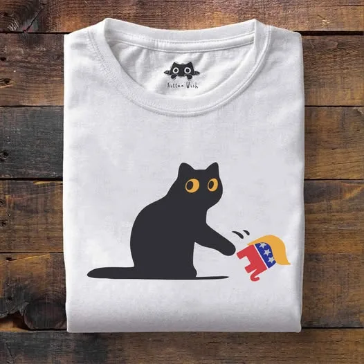 A folded white t-shirt on a wooden surface displays a black cat silhouette with yellow eyes, pooping on a red, white, and…