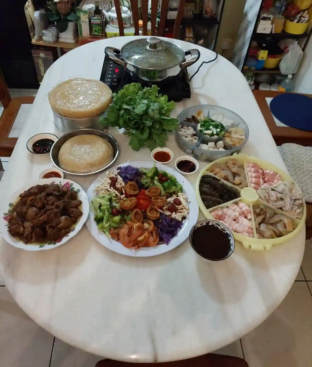 An overhead photo of a dining table laden with ingredients for a hot pot meal, including a covered pot on an electric…
