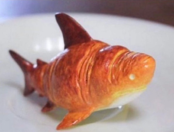 A golden-brown croissant shaped and baked to resemble a shark, complete with dorsal fin, tail, and gills, resting on a white…