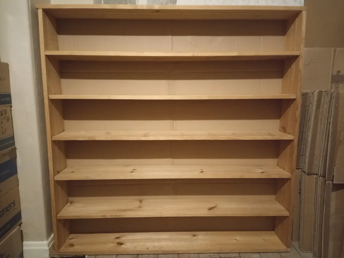 A large, empty wooden bookshelf with five shelves stands in a room surrounded by cardboard boxes. The shelves are bare…