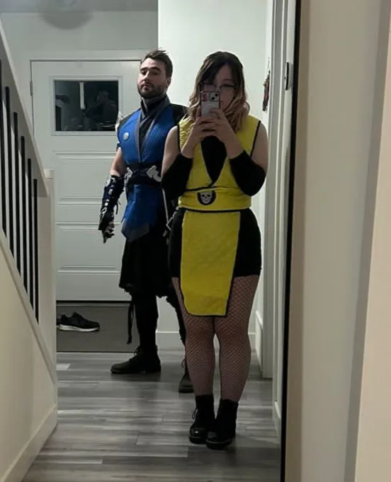 A person in a yellow and black maid outfit takes a mirror selfie while a man in a blue and black ninja costume stands behind…