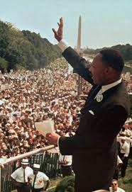 A historical photograph shows Martin Luther King Jr. standing on a platform, raising his right hand while addressing a…