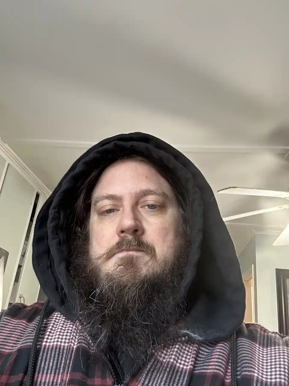 A man with a long beard and hood pulled up over his head stares directly at the camera with a neutral expression. He wears a…