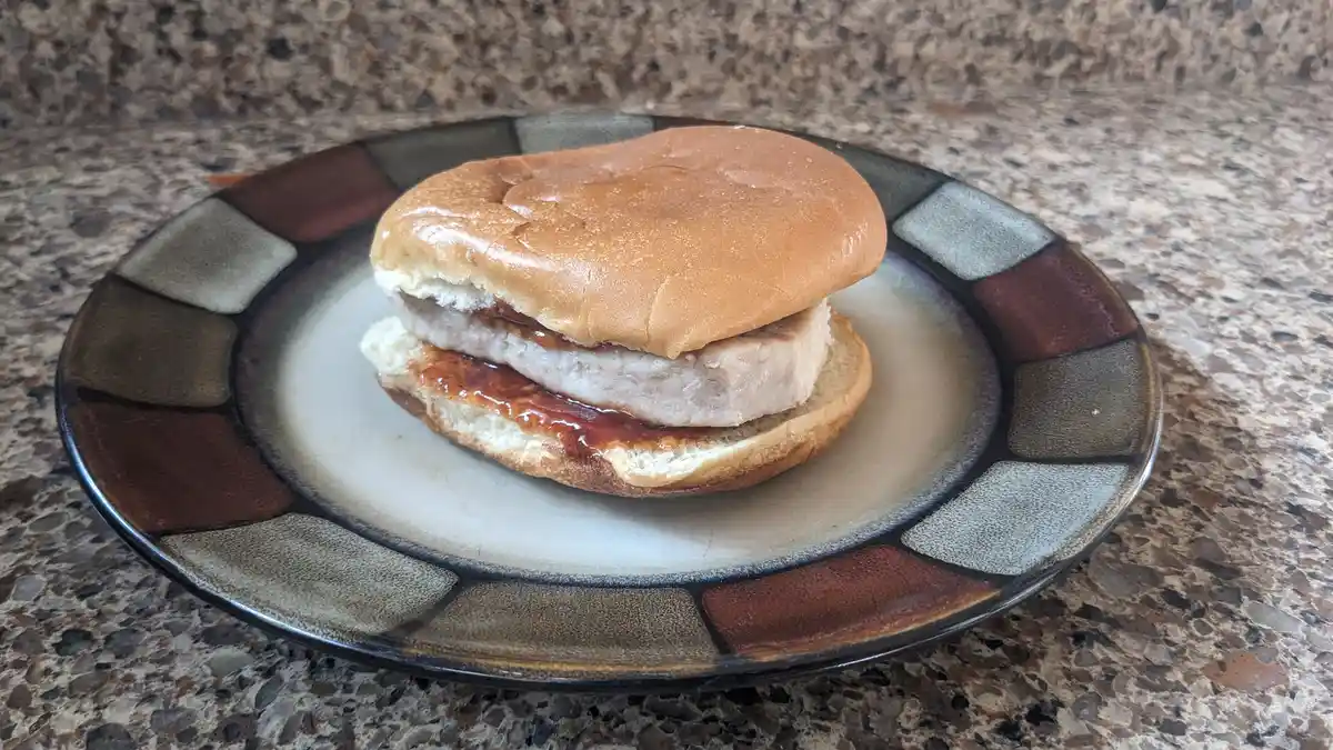 A simple sandwich made with a white bread bun, a single cooked chicken patty, and a layer of red sauce sits on a decorative…