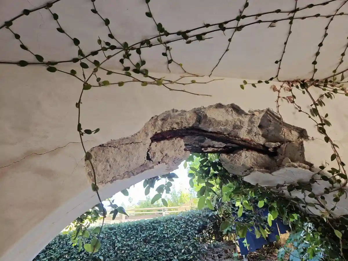 A close-up photo shows a broken, crumbling white plaster archway with vines growing over it, revealing a rusty metal beam…