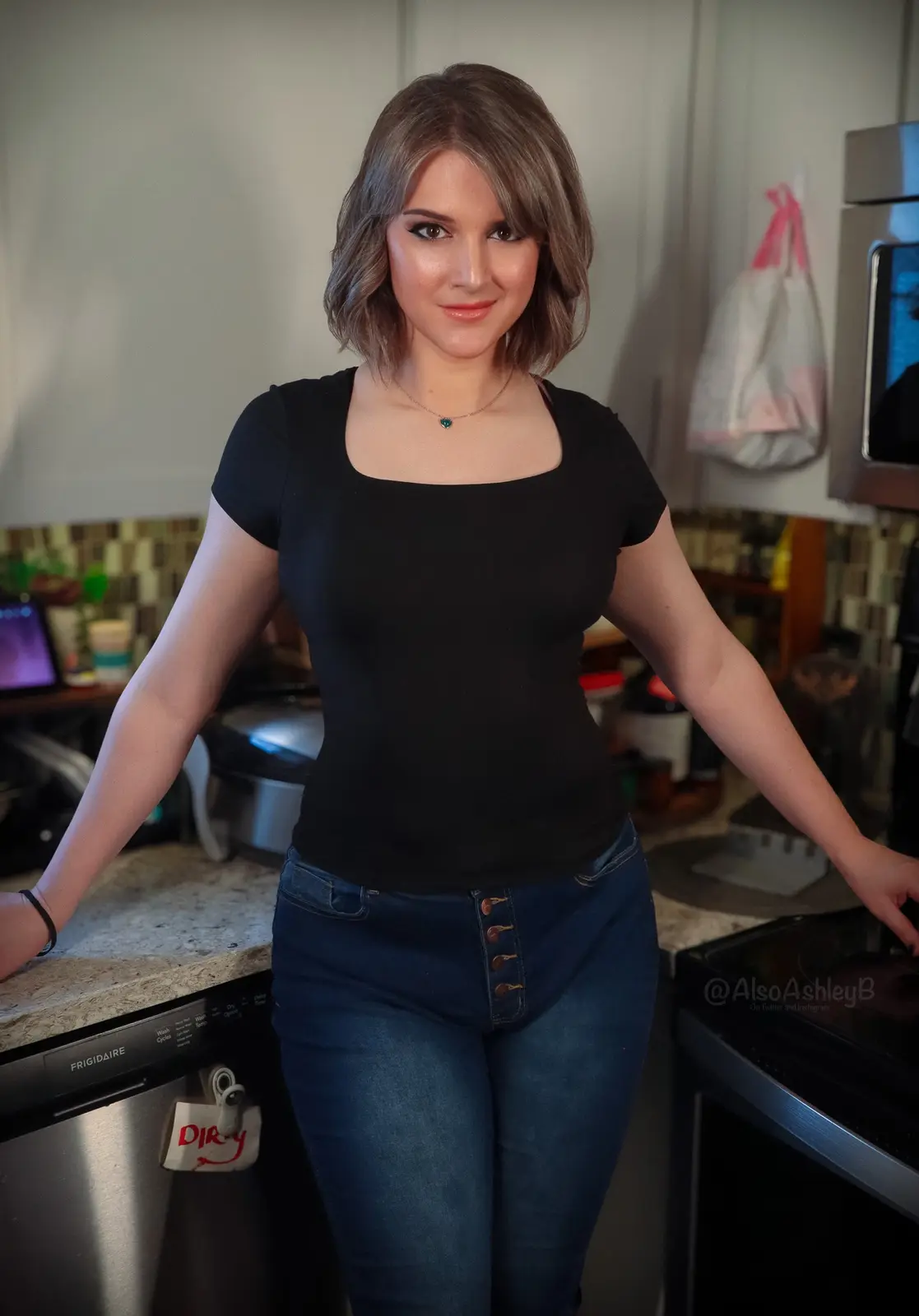 A woman stands confidently in a kitchen with her arms on countertops, wearing a black top and jeans. A small tag on the…