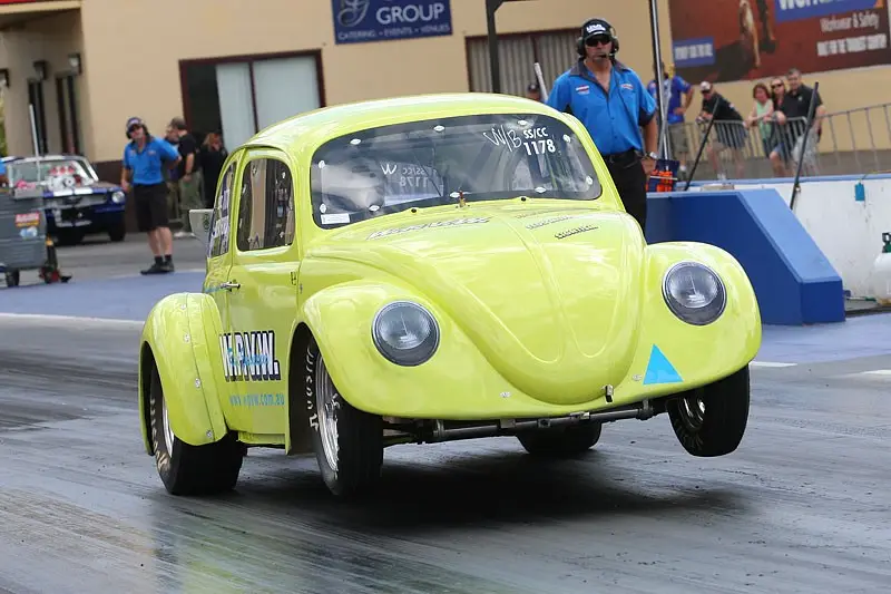 A lime green modified Volkswagen Beetle race car is airborne on a drag strip, with its front wheels off the ground. The car…
