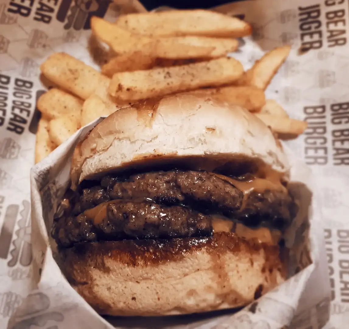 A close-up photo of a burger with multiple meat patties and cheese sauce, served with french fries on branded paper that…