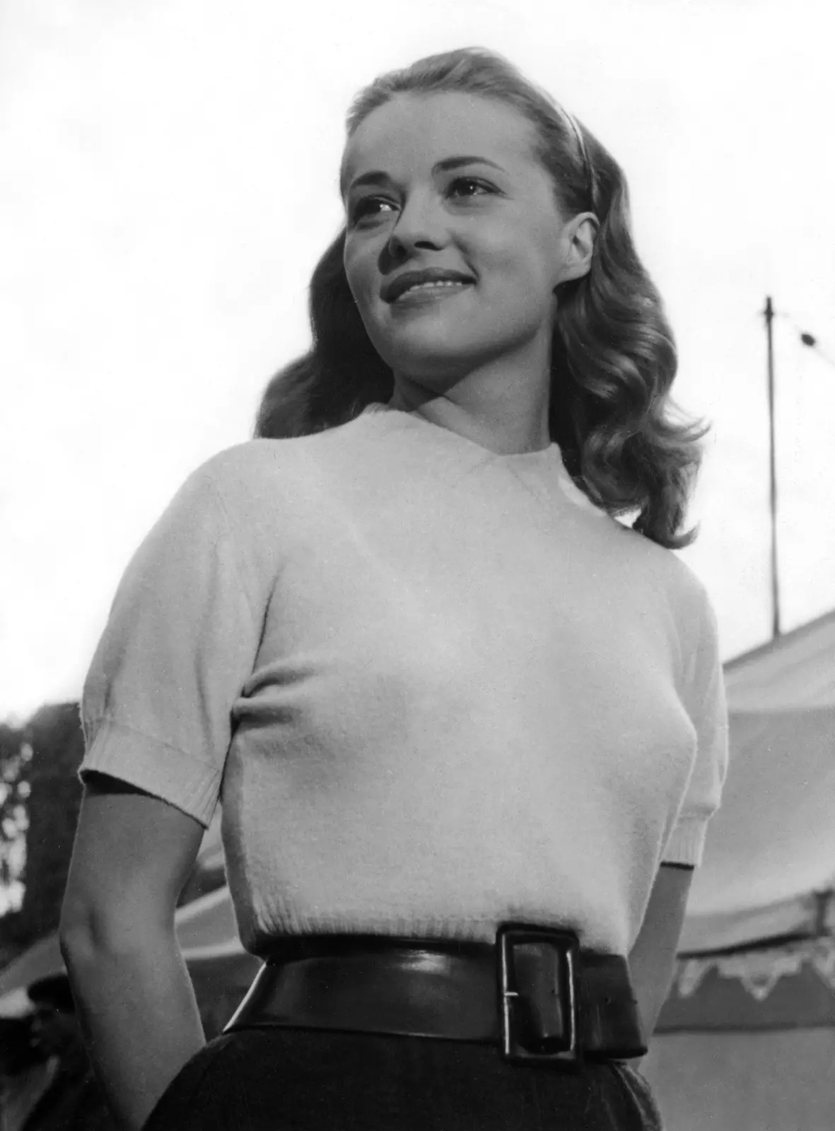 A black-and-white photo of a smiling woman in a light sweater and dark belt, looking upward with a pleasant expression. The…