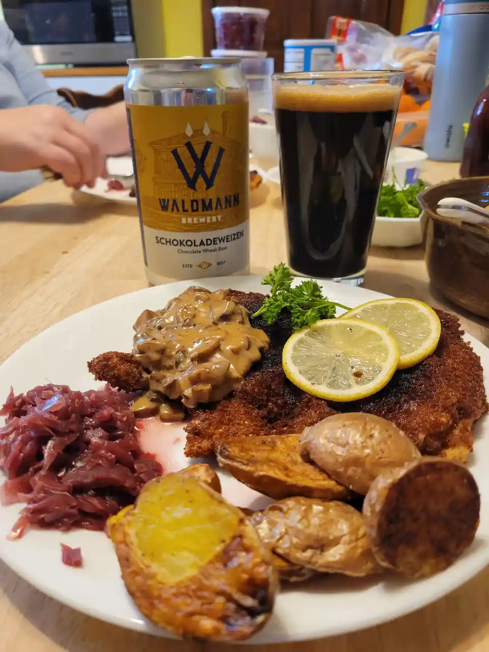 A plate of food including breaded meat with mushroom gravy, roasted potatoes, and red cabbage sits on a wooden table next to…