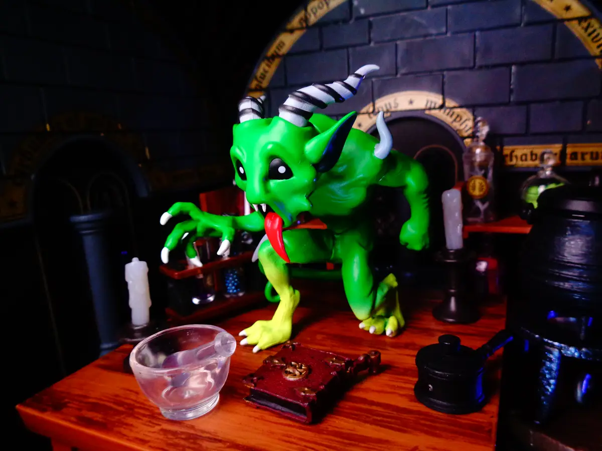 A green, horned demon figurine with a long red tongue is crouched on a wooden table in a dark, dungeon-like setting with…