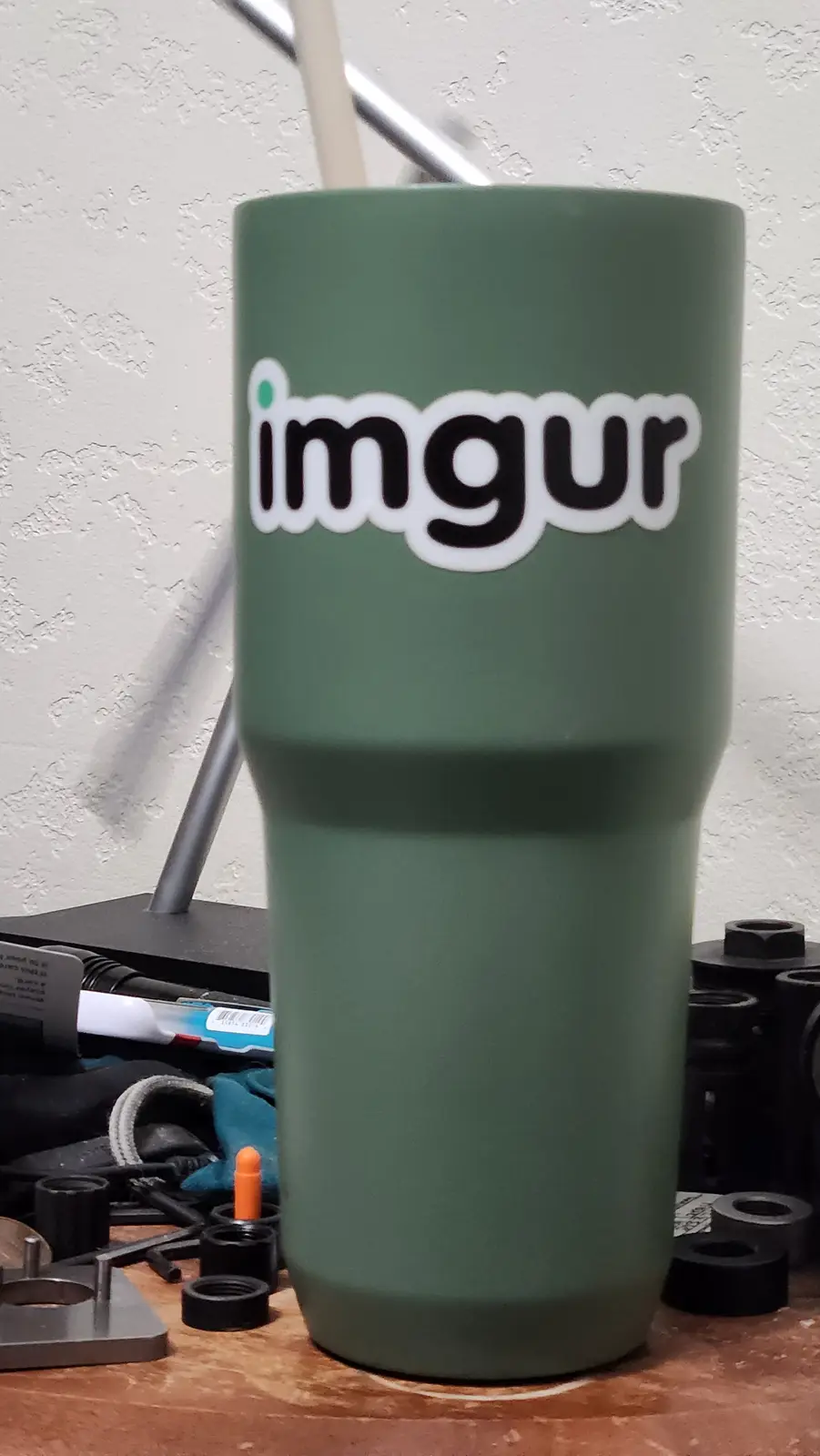 A matte green tumbler with the 'imgur' logo prominently displayed on its side sits on a cluttered workbench, with metal…