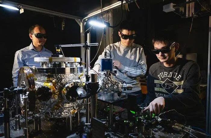 Three men in a lab wearing dark safety goggles stand around complex, shiny quantum computing equipment. The man on the right…