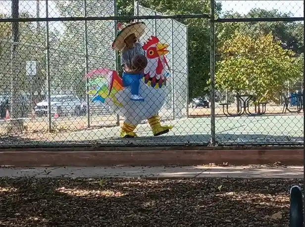 A person wearing a large, colorful rooster costume with a child sitting on their back, both viewed through a chain-link…