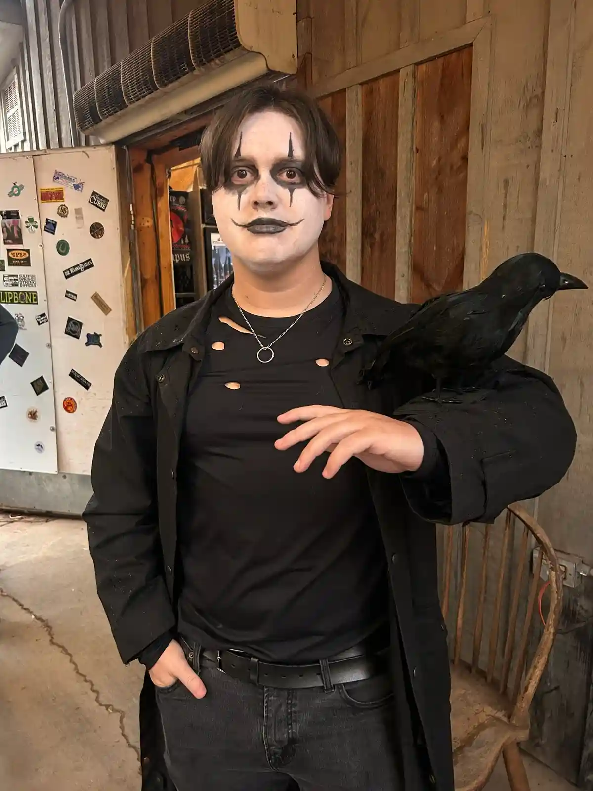A person dressed in a gothic vampire costume with white face paint, black eye makeup, and a black mustache, wearing a black…