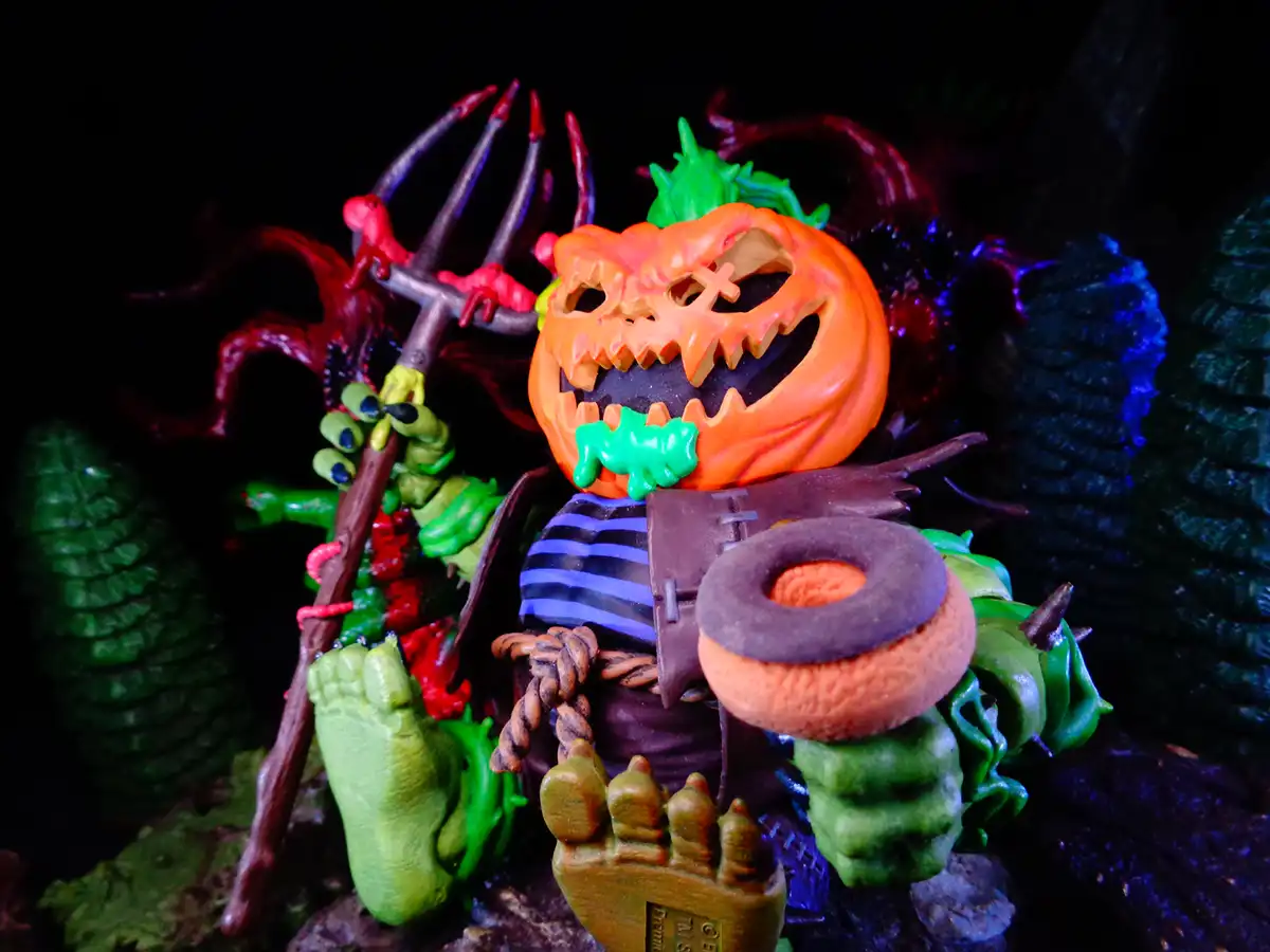 A Halloween figurine with a carved pumpkin head, green muscular body, and a donut on its head, holding a pitchfork. The text…