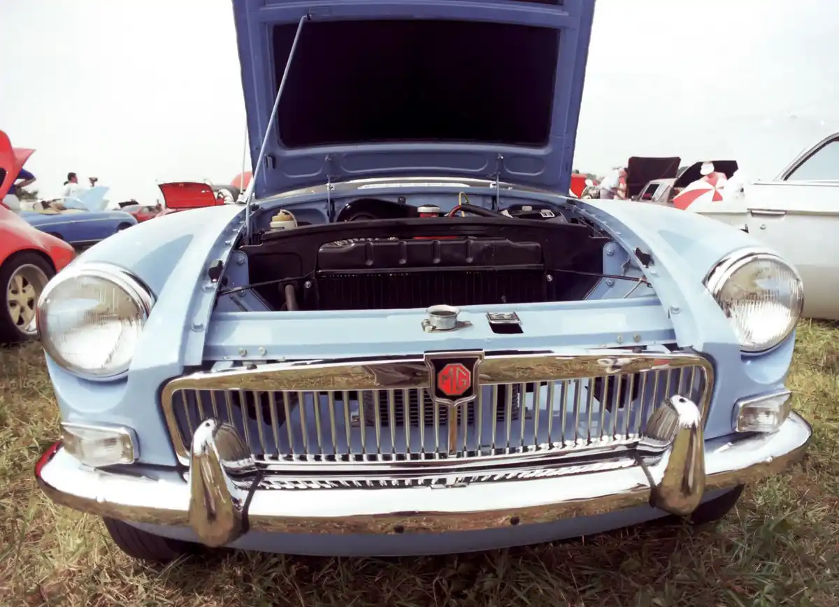 A light blue classic MG car with its hood open, revealing the engine bay, parked on grass at a car show with other vintage…