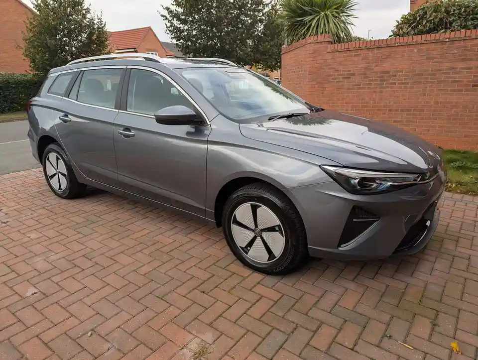 A grey MG electric car, likely an MG ZS EV, is parked on a brick driveway with a red brick wall and trees in the background…
