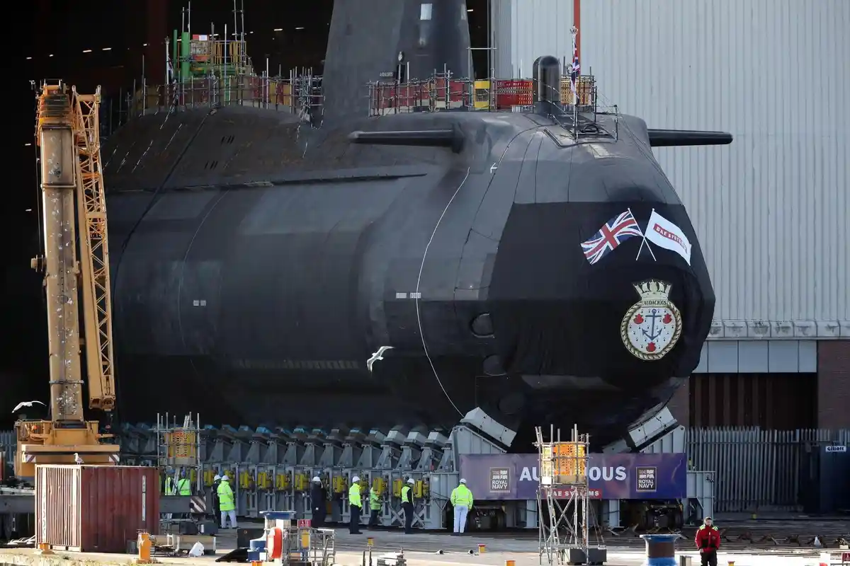 A large black submarine, identified as HMS Audacious by its crest and name on a banner, is shown in a shipyard with workers…