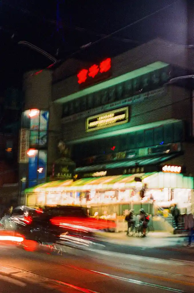 A long-exposure night photograph of a bustling Chinatown street, showing blurred streaks of car lights and indistinct…