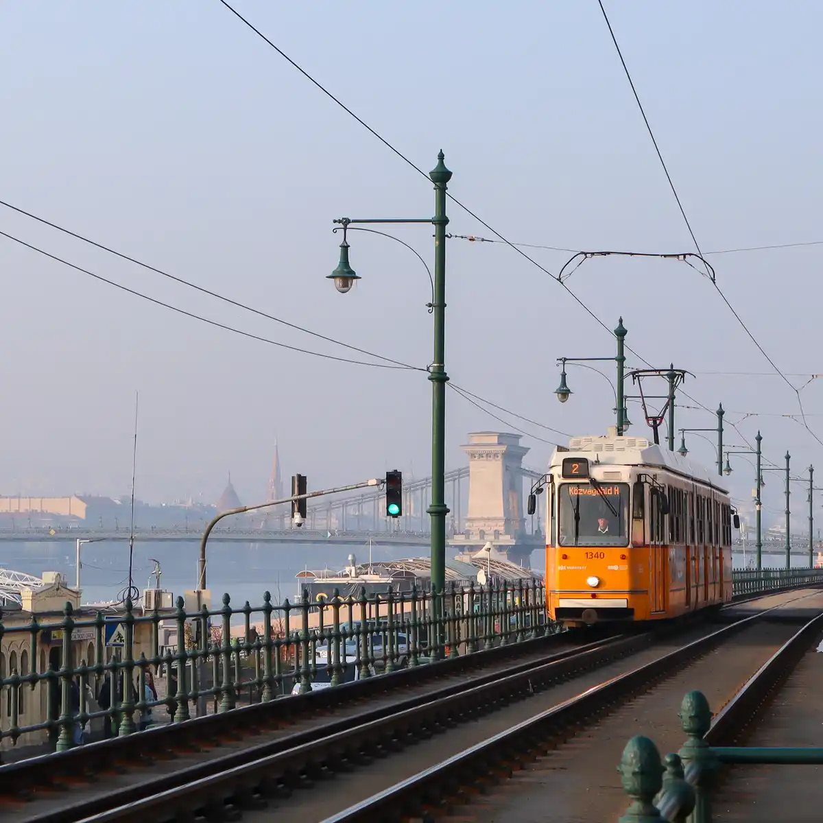 A yellow and white tram, numbered 1340 and heading to 'Közvágóhíd H', travels on tracks with a green traffic light…