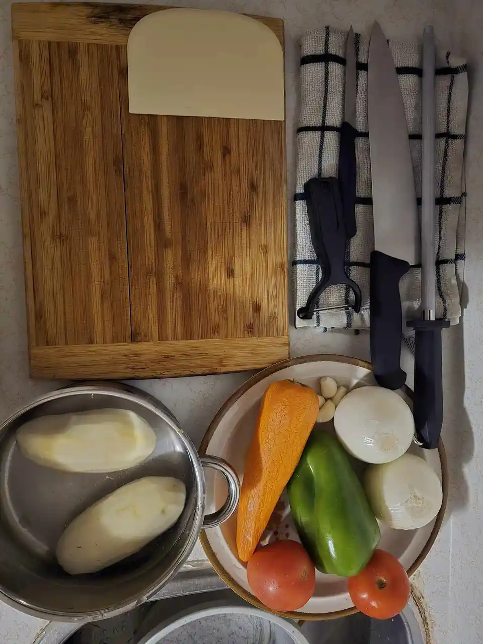An overhead shot of a kitchen counter with a wooden cutting board, a plate of vegetables including carrots, bell peppers…