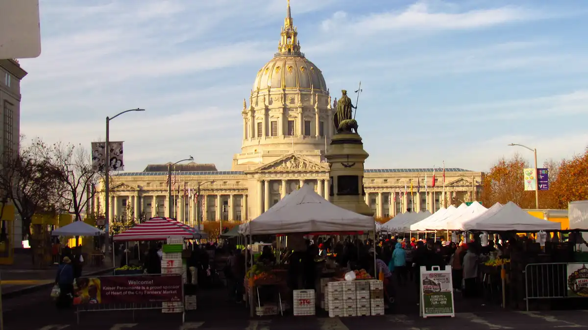 A bustling farmers market with white tents and produce crates is set in front of San Francisco City Hall, a grand…