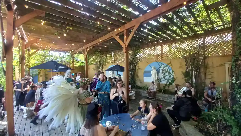 A group of people dressed in fantasy or historical costumes gather under a wooden pergola with string lights and ivy. A…