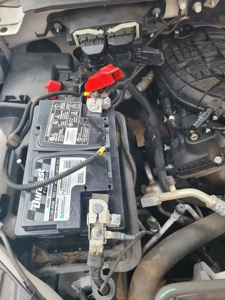 A close-up photo of a car's engine bay showing a Duracell AGM battery with red protective caps on its terminals, surrounded…