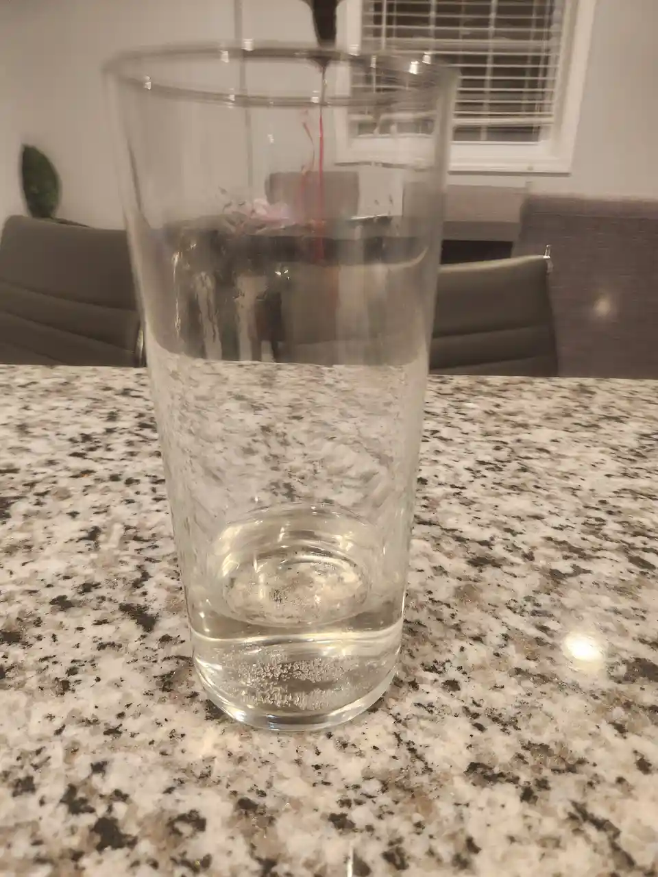 A clear drinking glass sits on a speckled granite countertop, containing only a small amount of water at the bottom. A thin…