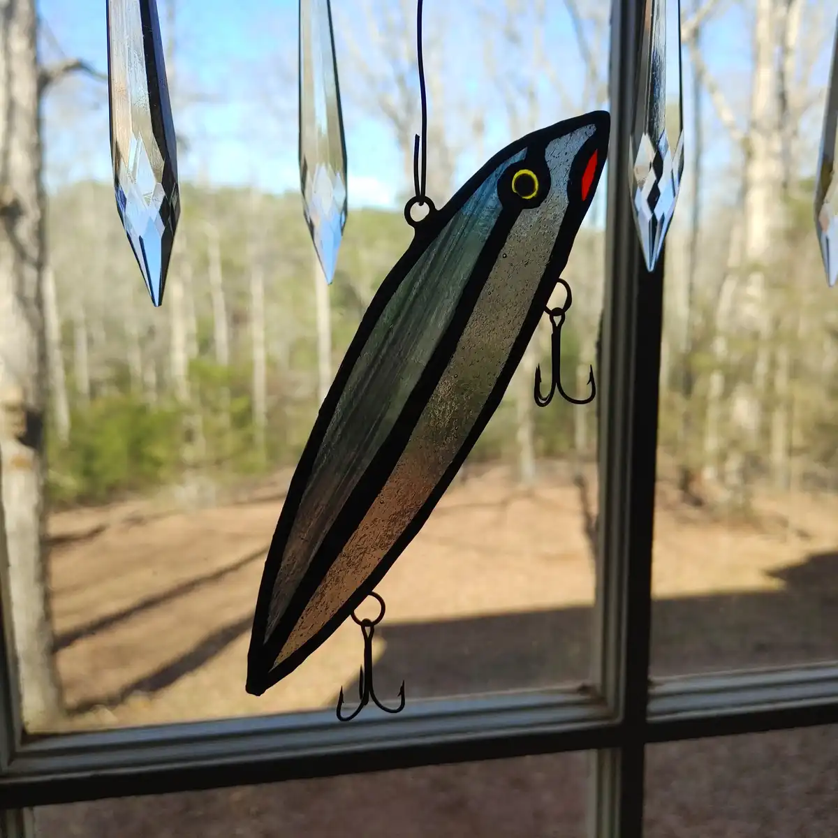 A stained glass window hanging shaped like a fishing lure with two hooks, suspended in front of a window with a forest view…