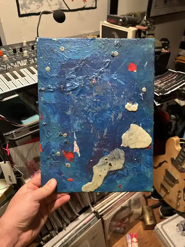 A hand holds up a textured, abstract painting dominated by blue with white and red paint splatters and patches, set against…