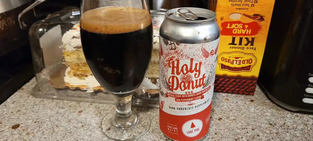 A can of 'Holy Donut' dark chocolate peppermint imperial stout sits next to a glass of the dark beer, with a slice of cake…