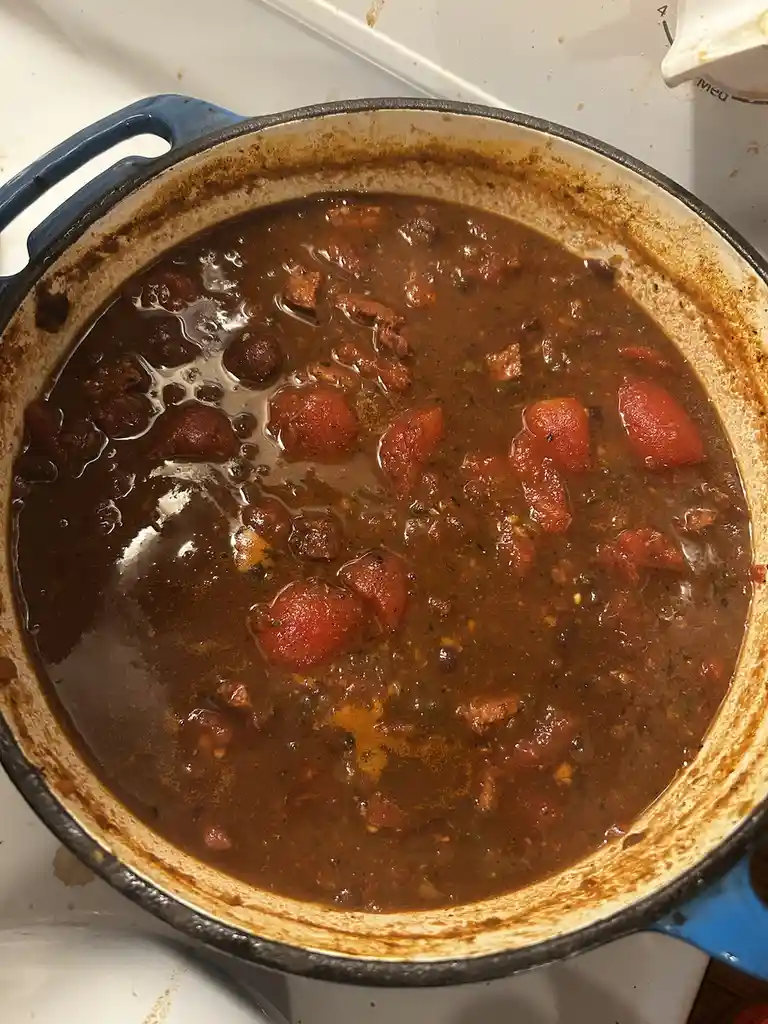 A close-up photo shows a blue enameled cast iron Dutch oven on a stovetop, filled with a dark brown stew or chili containing…