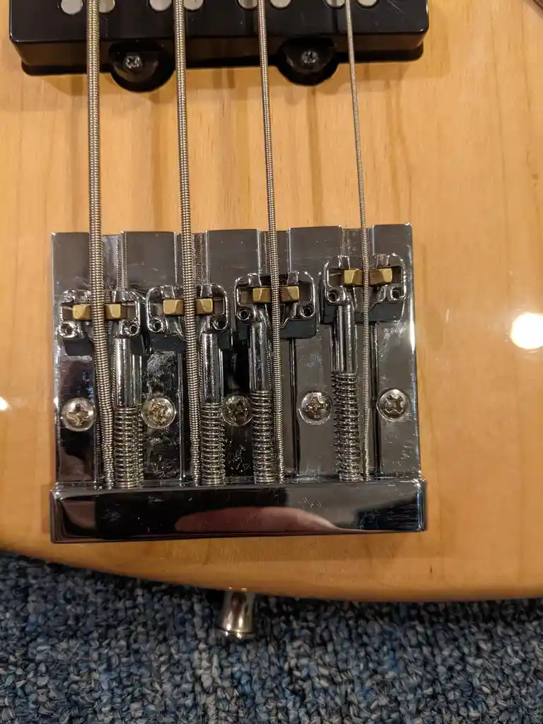 A close-up photo of a bass guitar's chrome bridge with four strings attached, set against a light wood body and blue carpet…