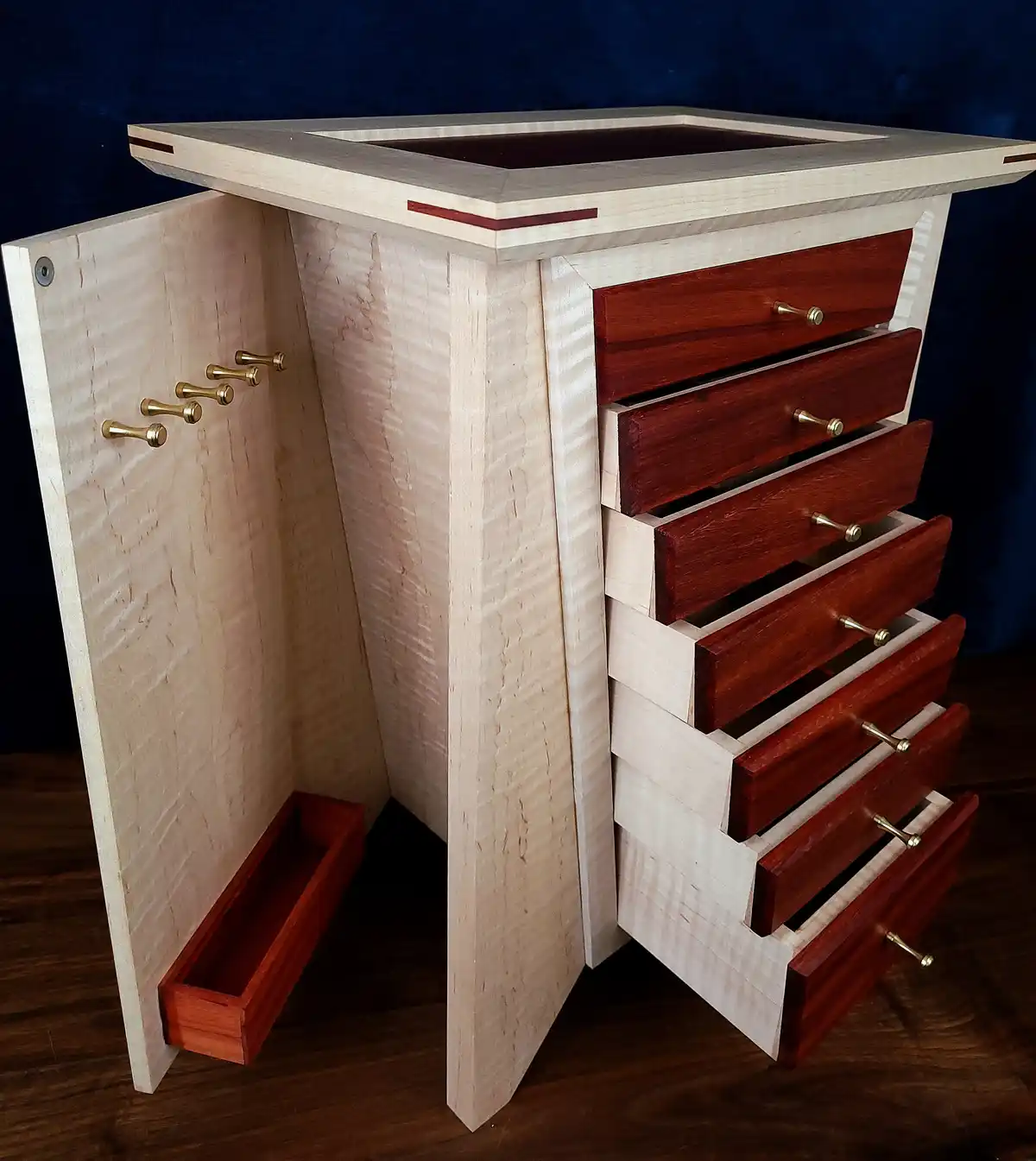 A custom wooden jewelry box with a light-colored wood frame and dark reddish-brown drawers, partially open to reveal…