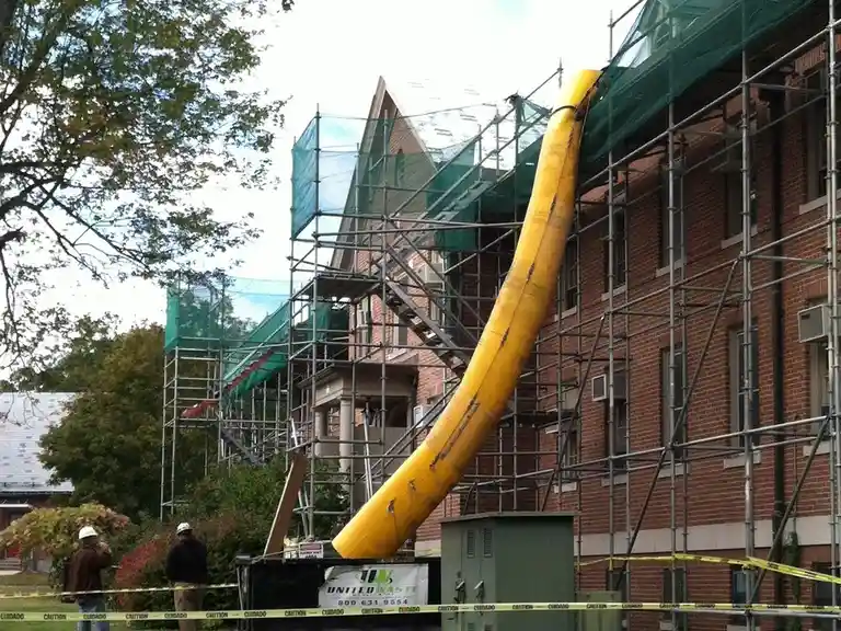 a large yellow plastic slide is attached to the side of a brick building covered in metal scaffolding and green safety…