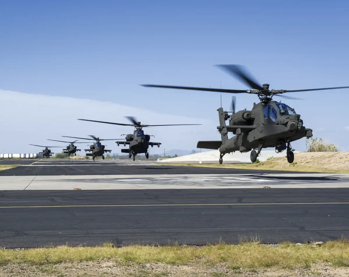 A line of military Apache helicopters takes off in formation from an airstrip under a clear blue sky. The closest helicopter…