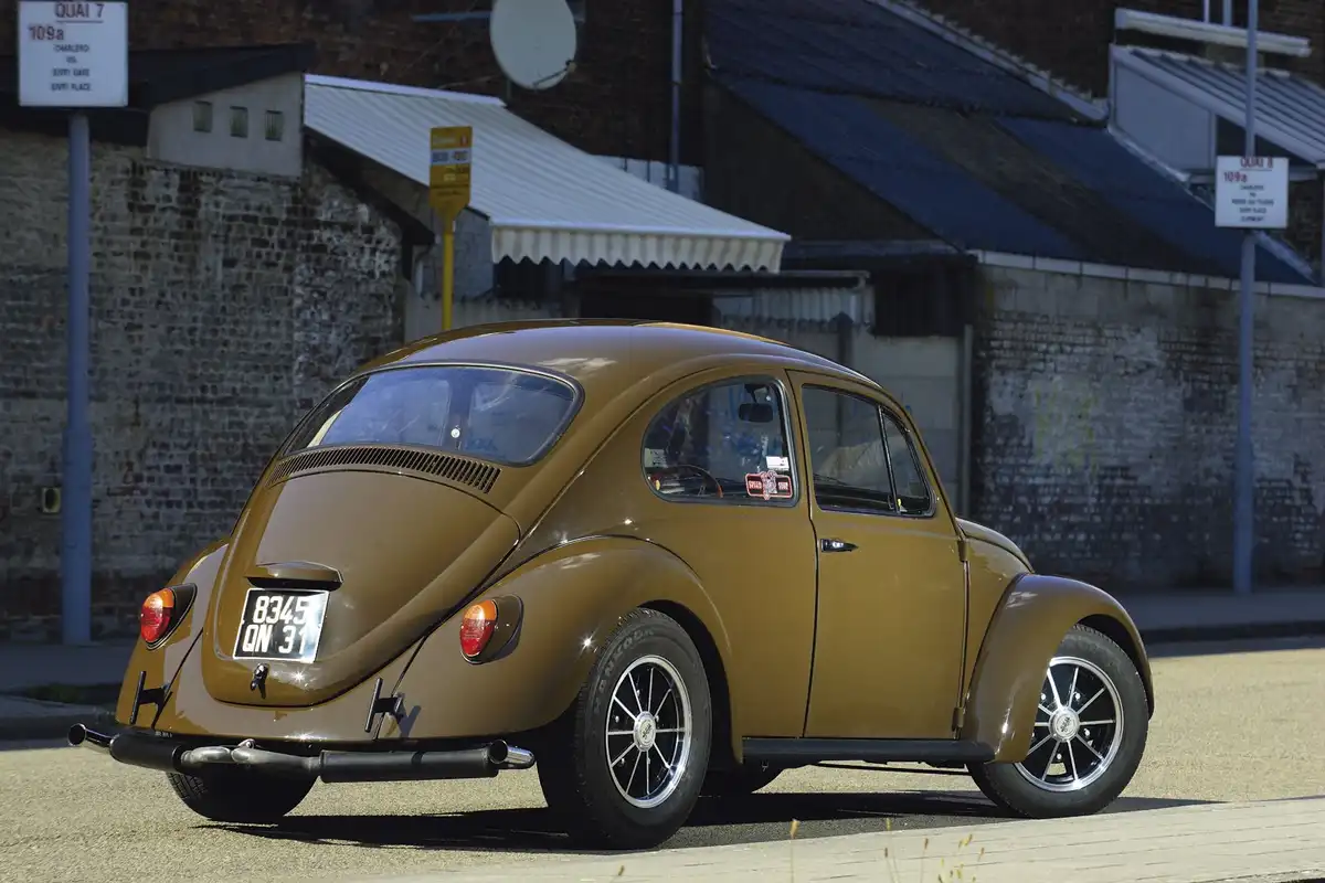 A brown vintage Volkswagen Beetle is parked on a street next to brick buildings with signs for 'QUAI 7' and 'QUAI 8'. The…