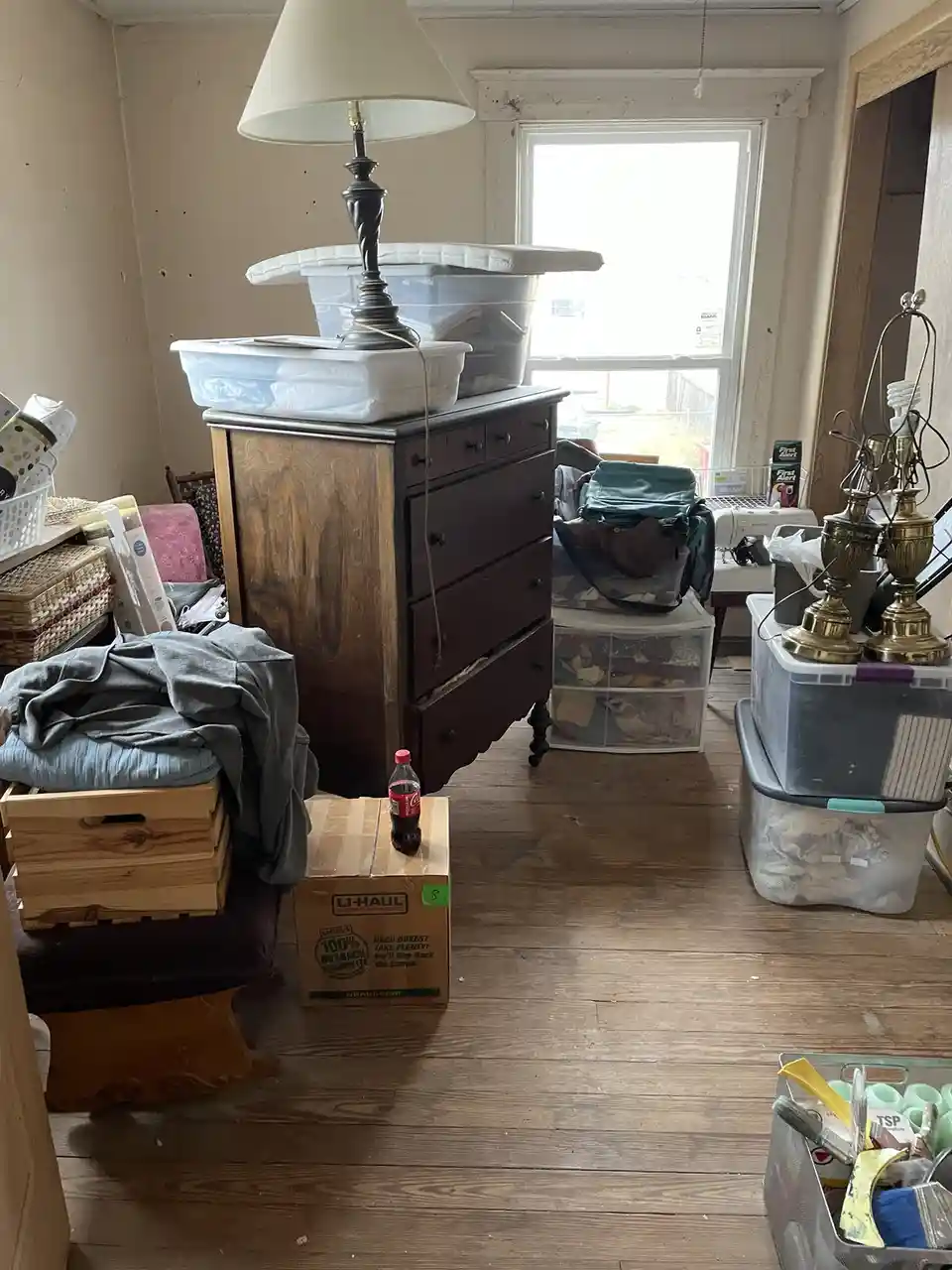 A cluttered room filled with furniture, storage bins, and boxes, including a U-Haul box with a single Coca-Cola bottle on…