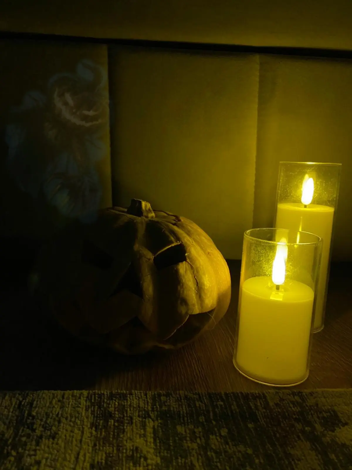 A dimly lit scene shows a carved jack-o'-lantern and two lit candles in glass holders on a wooden surface. The warm yellow…