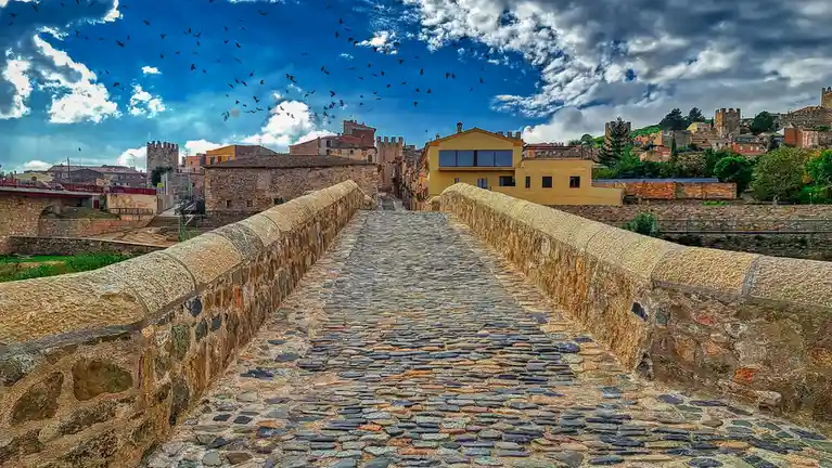 A wide-angle photo shows a cobblestone stone bridge leading toward a historic town with castle-like buildings under a…